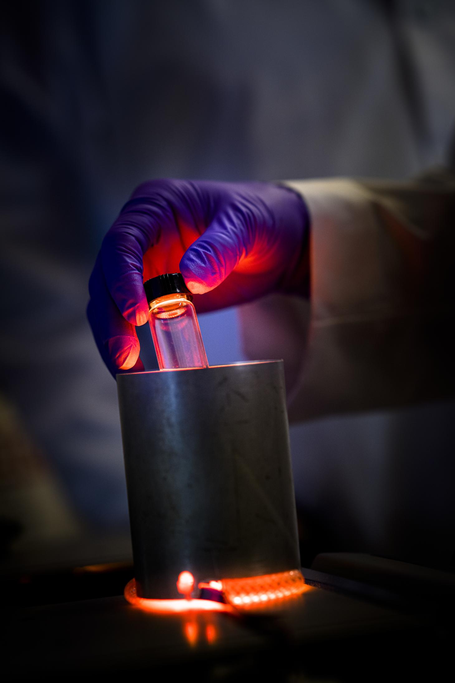 A gloved hand holding a beaker above a heater in a lab.