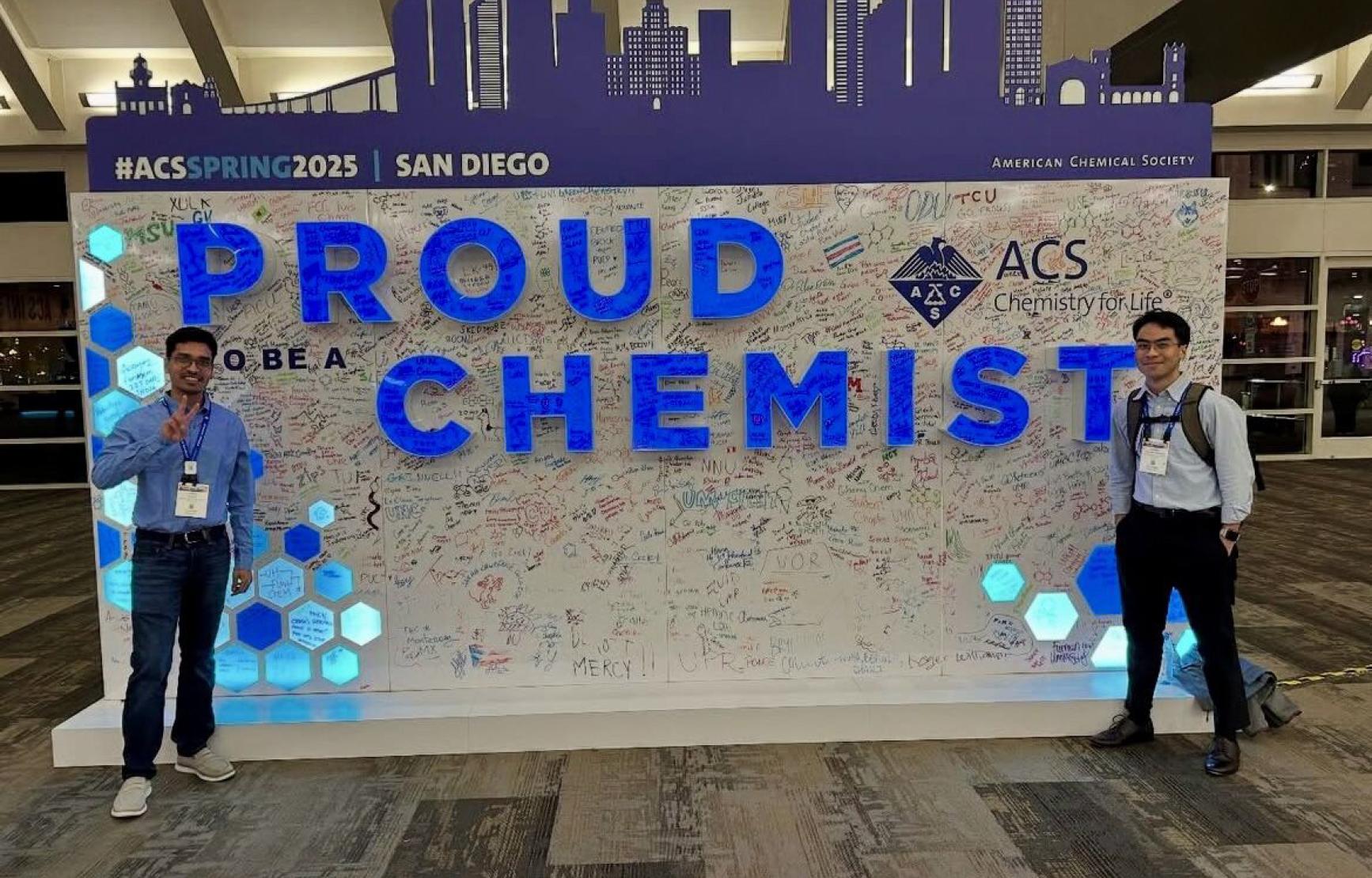 Two graduate student flank a large sign at the 2025 American Chemical Society conference that says "Proud to be a Chemist"