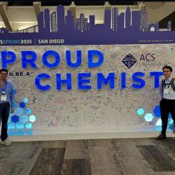 Two graduate student flank a large sign at the 2025 American Chemical Society conference that says "Proud to be a Chemist"