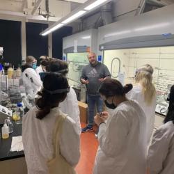 A group of students in lab coats stand around a professor in a laboratory.