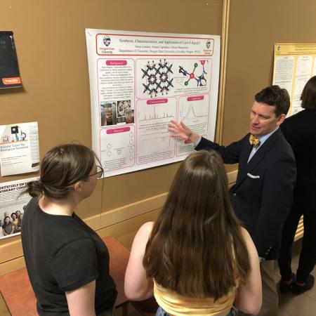 A student shows a group of people a research poster hanging on a wall. 