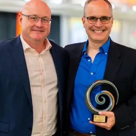 Two men stand wearing suits. One of them is holding an award.
