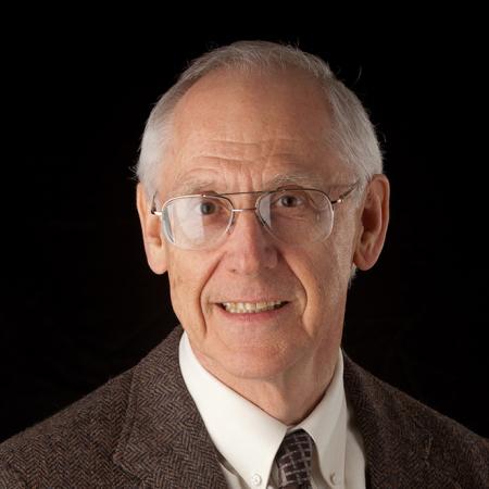 A man's headshot against a black background. The man has gray hair and wears glasses with a brown suit jacket and tie.