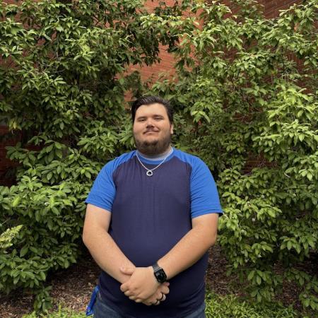 A smiling man stands outside the chemistry building with hands clasped