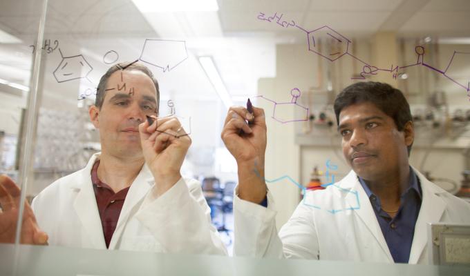 Two chemistry faculty members write equations on a glass board.