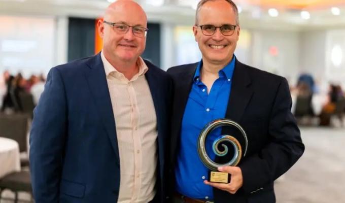 Two men stand wearing suits. One of them is holding an award.