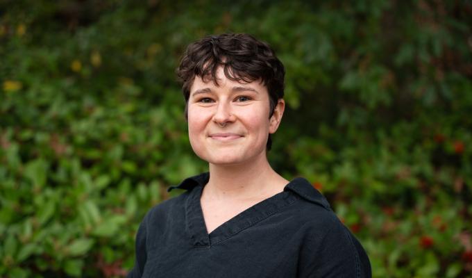 Sophia Bailey smiles in front of Oregon greenery. She wears a black button-up shirt and has short cropped, curly, dark hair. 