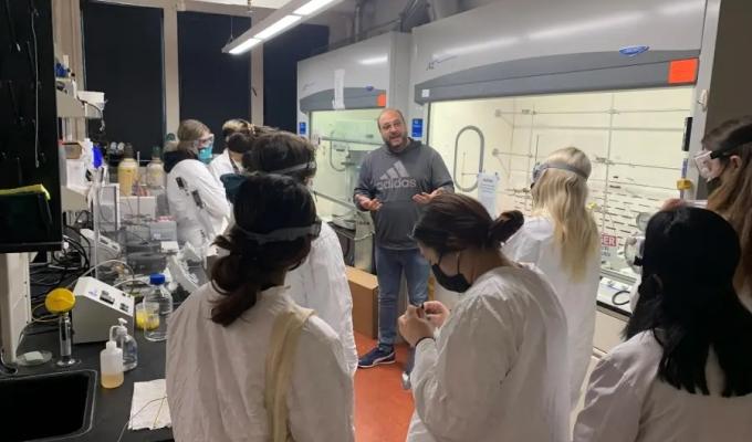 A group of students in lab coats stand around a professor in a laboratory.