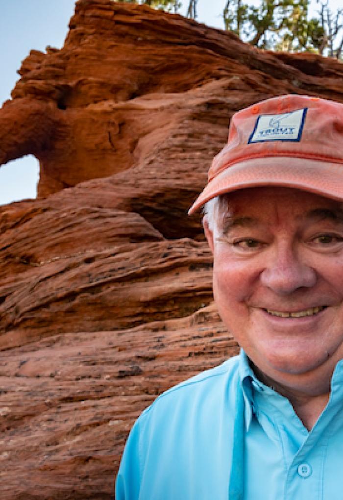 Chuck Armstrong, '66 where's a light blue collared shirt and a salmon colored baseball cap. He stands in front of rock formations on a recreational outing. 