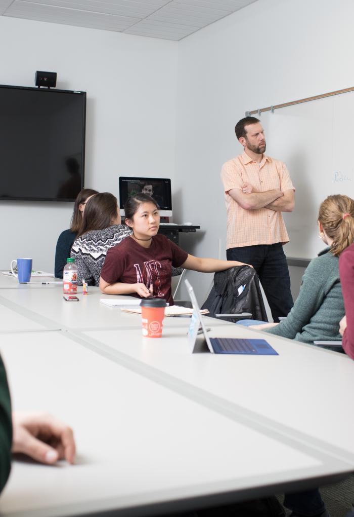 Students sit around a table and look towards an instructor in a salmon colored shirt.