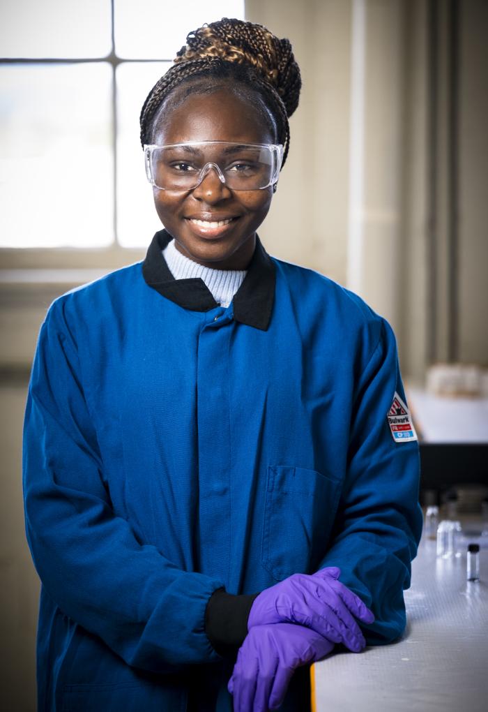 A woman in a lab coat, goggles and gloves poses for a headshot.