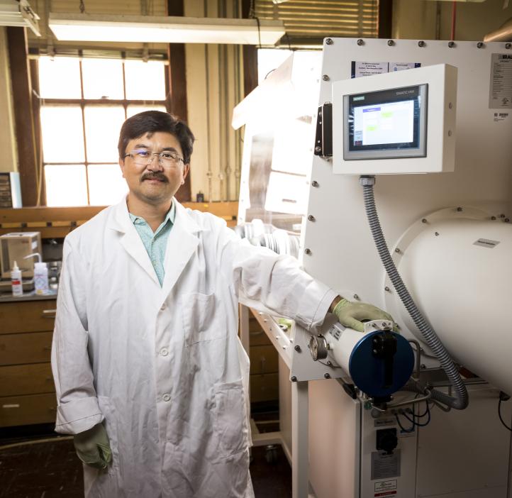 A man in a lab coat stands in front of a white machine used for battery science.