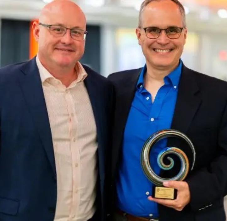 Two men stand wearing suits. One of them is holding an award.