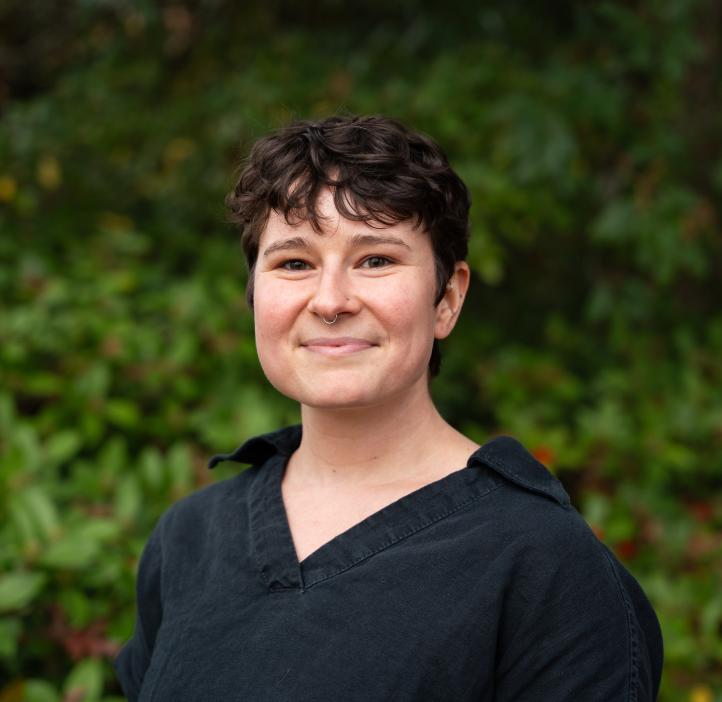 Sophia Bailey smiles in front of Oregon greenery. She wears a black button-up shirt and has short cropped, curly, dark hair. 