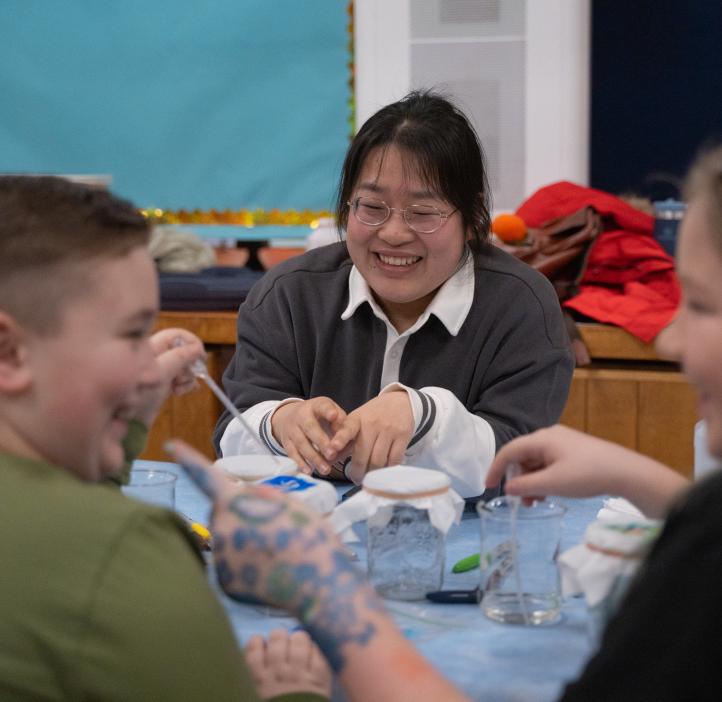 An OSU student laughs at the two children across the table from her engaged in shenanigans. Both children are laughing and one is pointing at the other. The experiment is on the table between them.