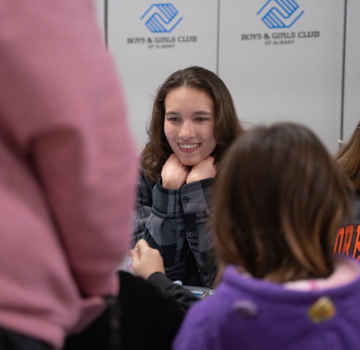 An OSU student has their hands tucked under their chin and their elbows on the table. She is smiling widely on the children across the table from her.