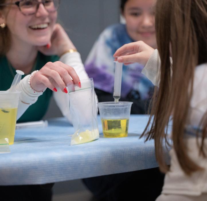 An OSU student smiles widely at the child in front of them. She holds a baggie of yellow liquid that the child is using a syringe to extract and place into a cup in front of her.
