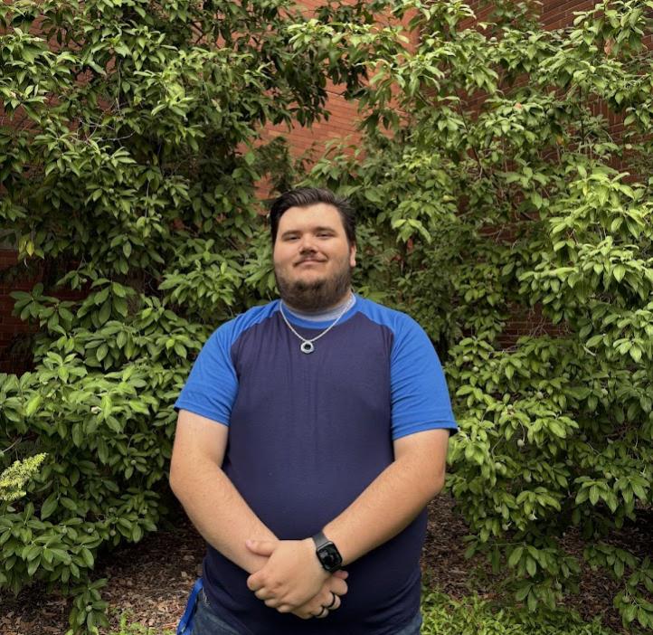 A smiling man stands outside the chemistry building with hands clasped